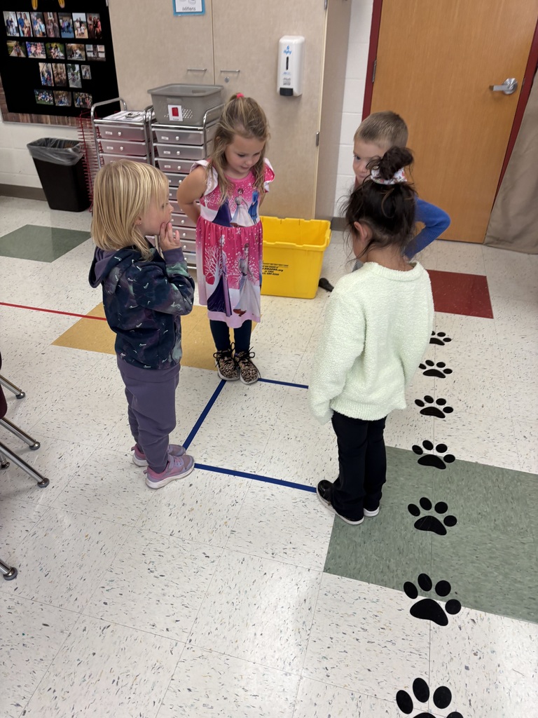 Four students stand in a small square outlined with blue tape on the classroom floor, looking down as they participate in a hands-on learning activity.