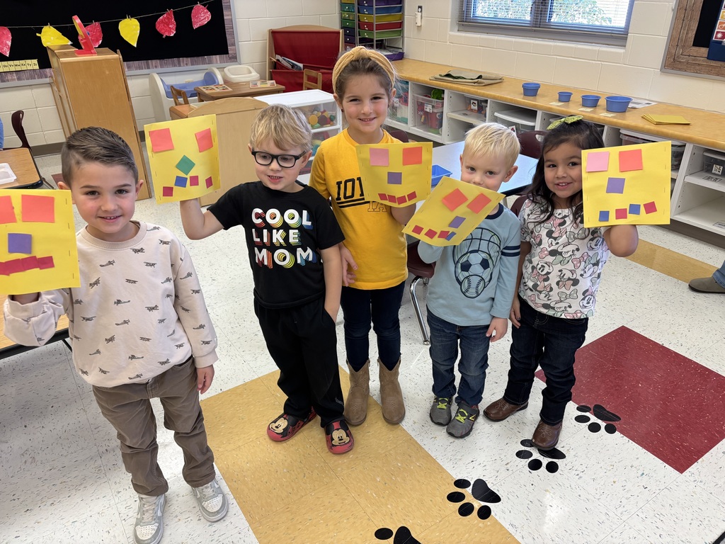 Five young students stand in a row, proudly holding up yellow papers decorated with colorful geometric shapes.