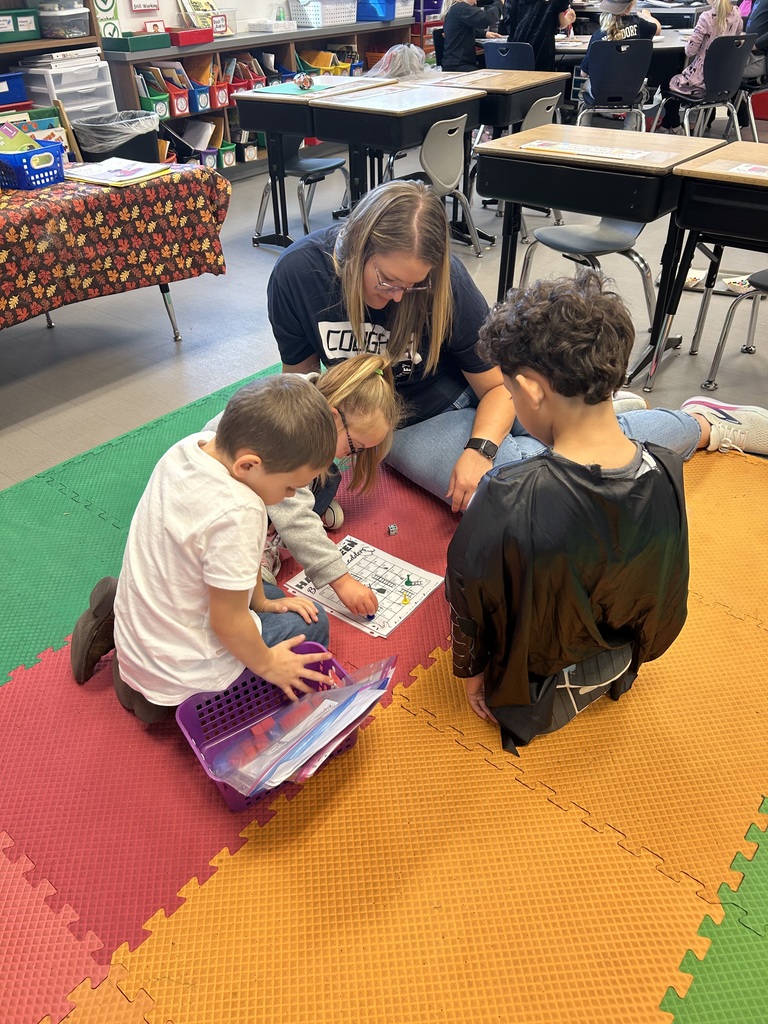 A teacher sits on colorful foam mats with three young students, helping them play an educational board game with dice and small pieces