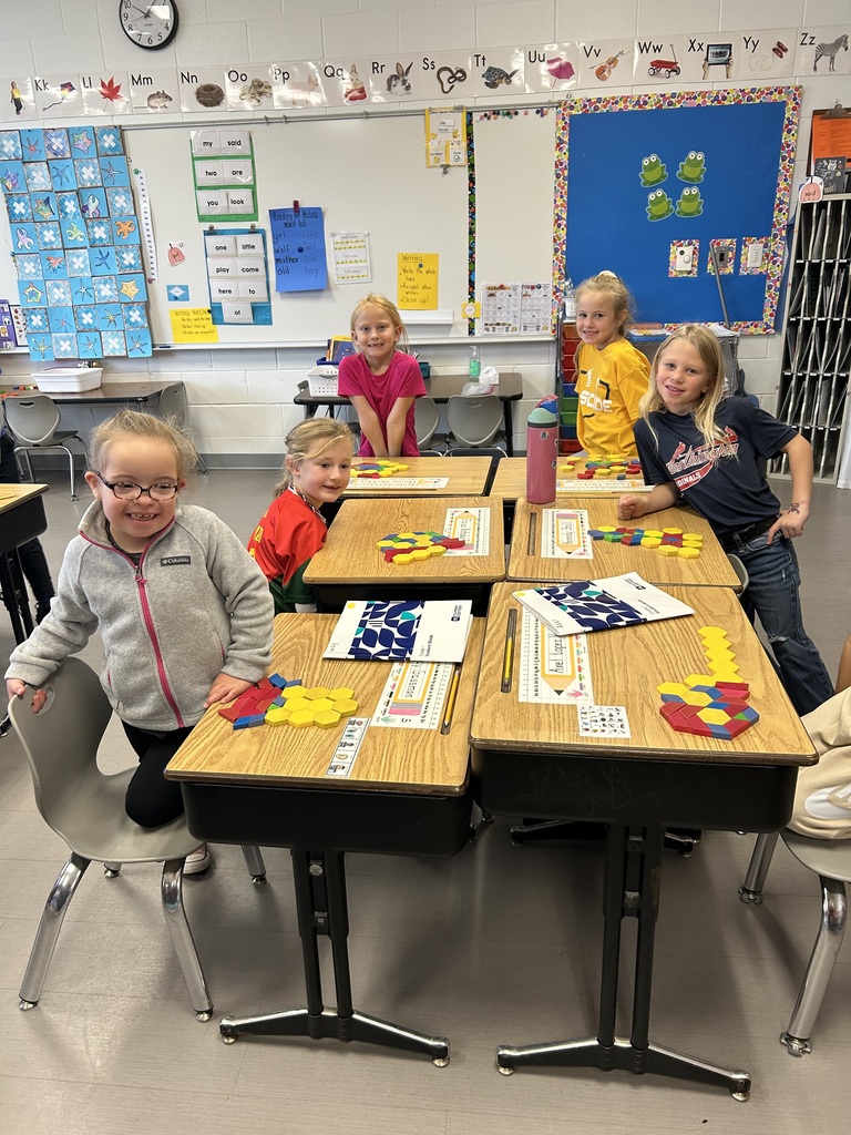 Five students sit and stand around grouped desks, smiling as they work with colorful pattern blocks to create geometric shapes.