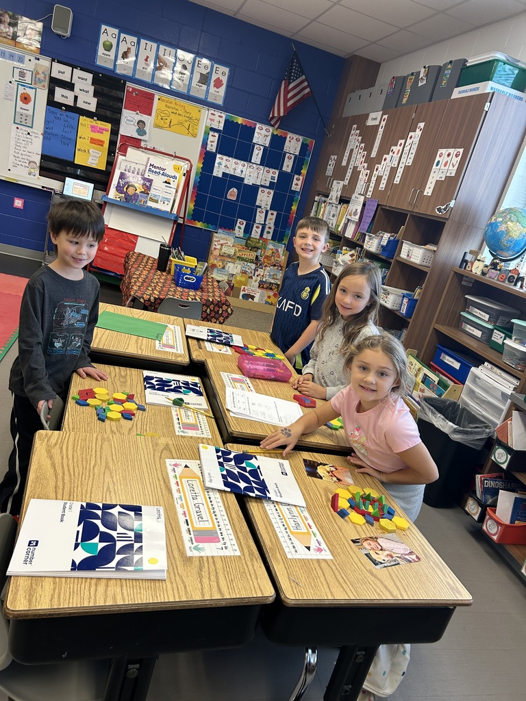 Four students stand at their desks, smiling as they work with colorful pattern blocks on math activities.
