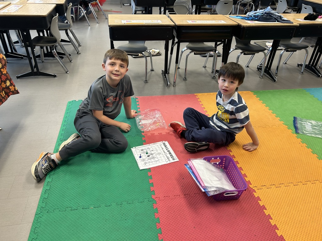 Two young students sit on colorful foam mats, smiling at the camera.