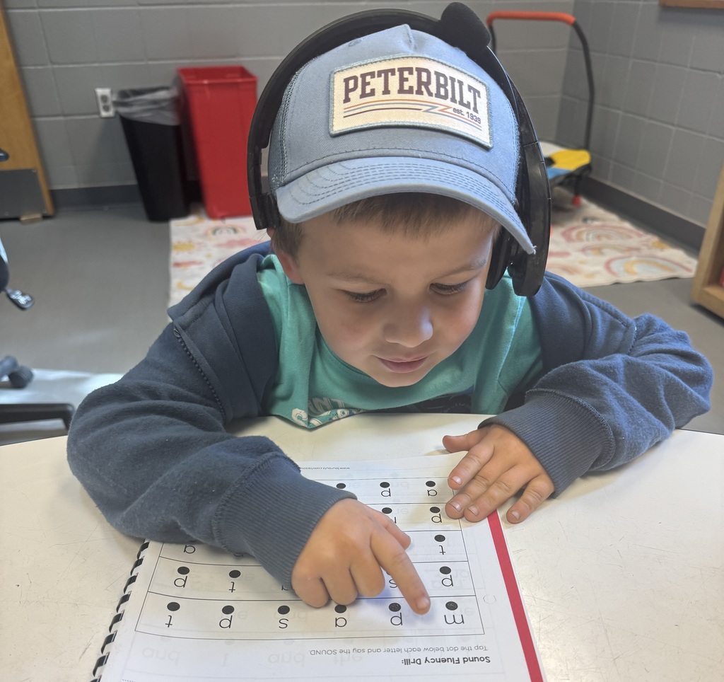 A young boy wearing a blue “Peterbilt” hat and headphones works on a phonics worksheet. He points to a row of letters on the page while leaning over the table.