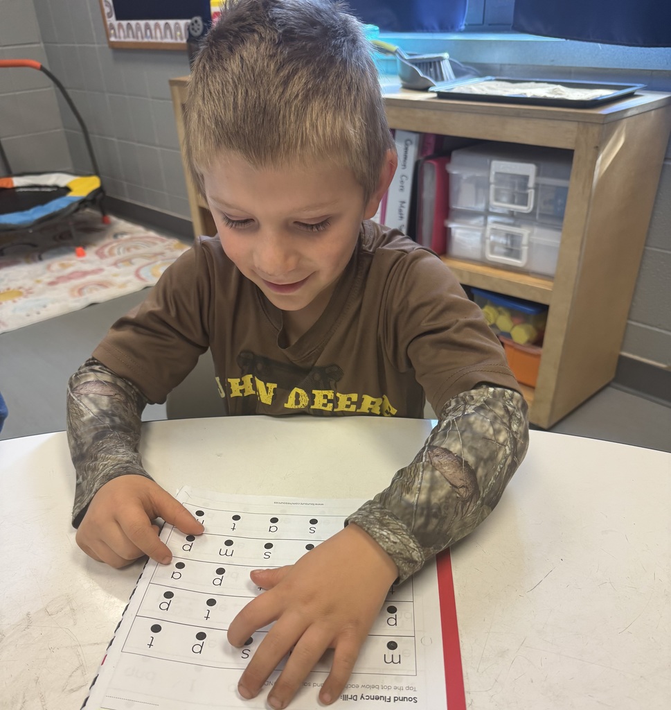 A young boy sits at a table working on a phonics worksheet. He points to letters on the page while smiling and concentrating. He wears a brown “John Deere” shirt and camouflage sleeves.