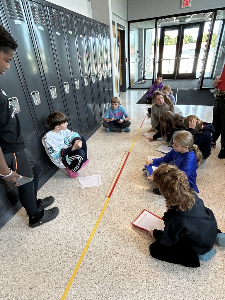 Students sit in a hallway near a row of black lockers, writing on worksheets clipped to clipboards.