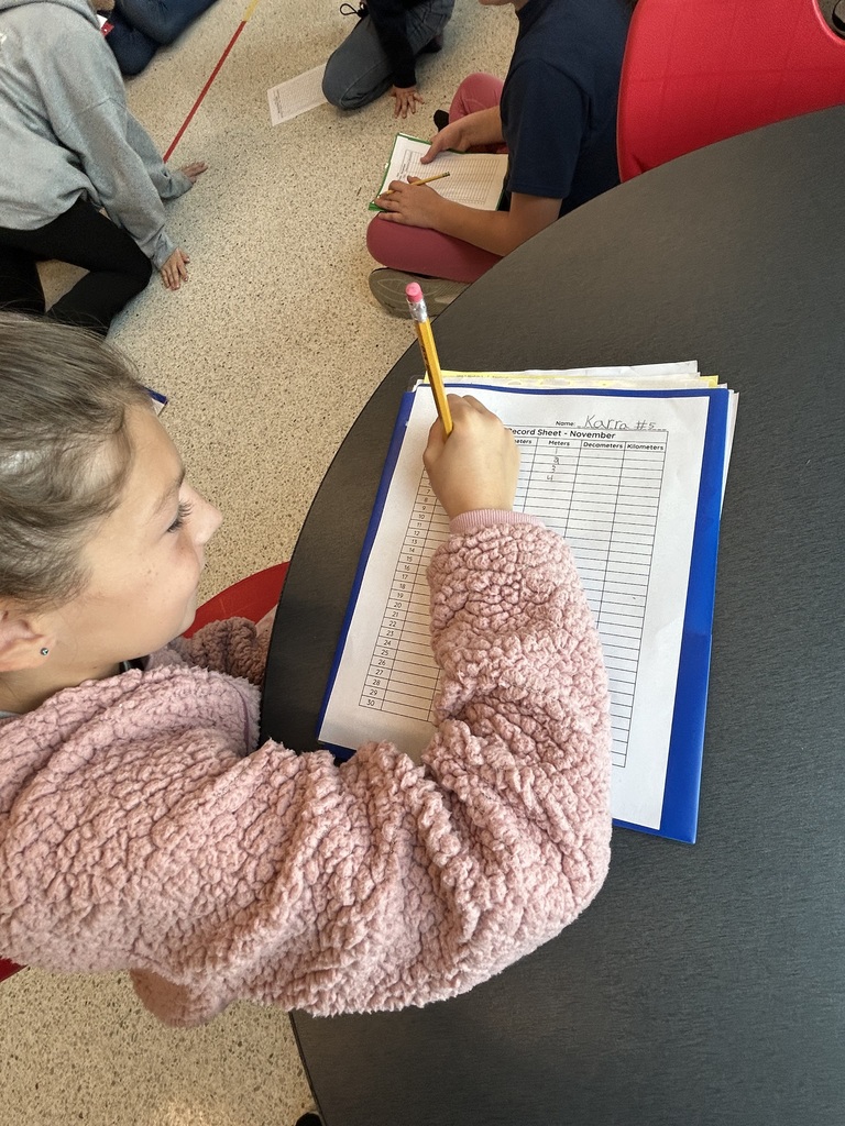 A student wearing a pink fuzzy jacket sits at a round table and writes on a November record sheet clipped to a blue clipboard.