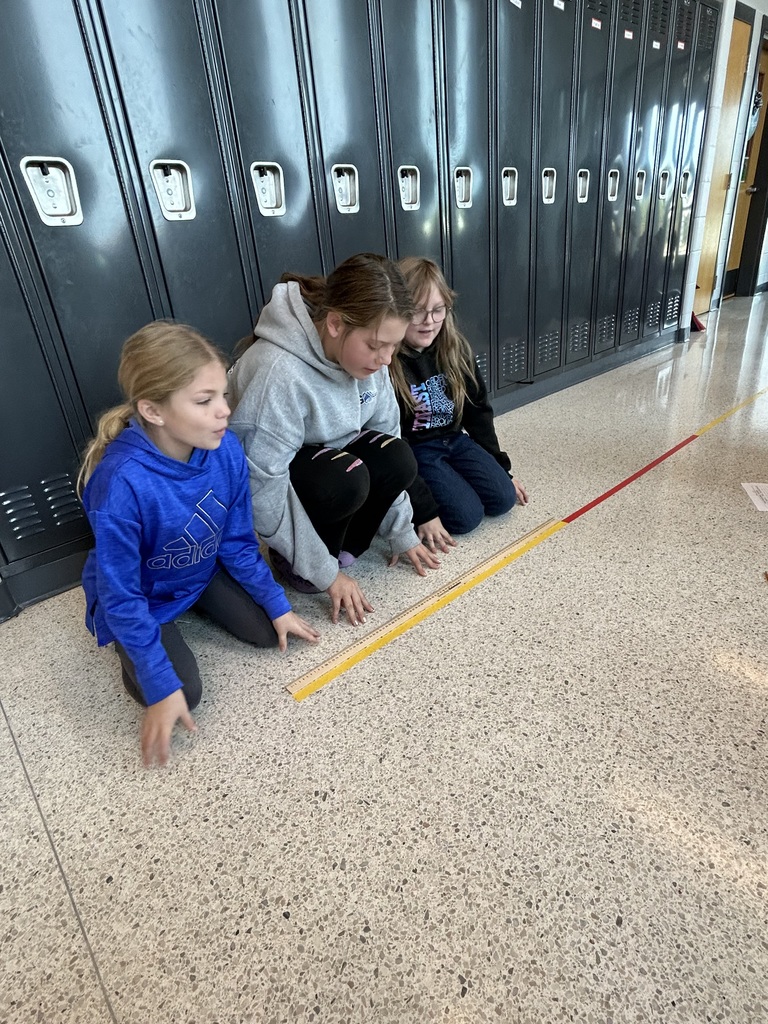 Three students kneel on the hallway floor next to black lockers, measuring along a yardstick placed on the ground.