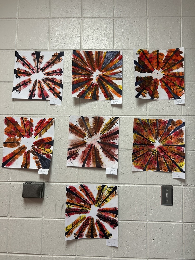 Seven pieces of student-created radial artwork in red, orange, yellow, and black tones displayed on a white tiled wall, each labeled with a student’s name.