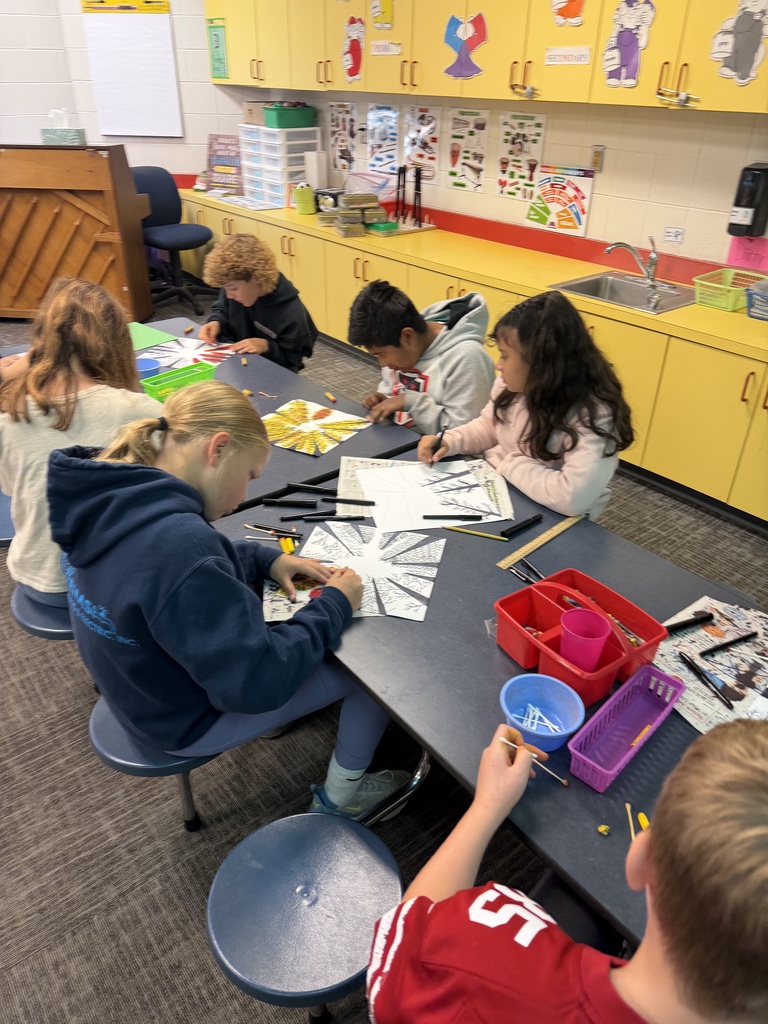 Students seated around a table in an art room working on black-and-white radial art designs, adding color with crayons and tools from small plastic bins.