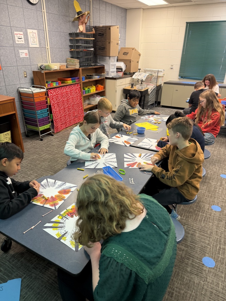 Students sitting at a long table in an art room, coloring fall-themed radial art designs with crayons and cotton swabs.