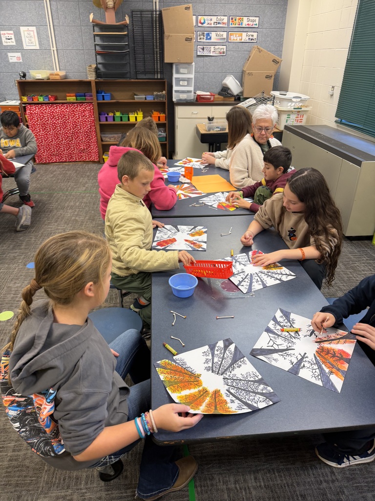 A group of students seated at a rectangular table creating fall-themed radial artwork, with an adult assisting a pair of students on the right.