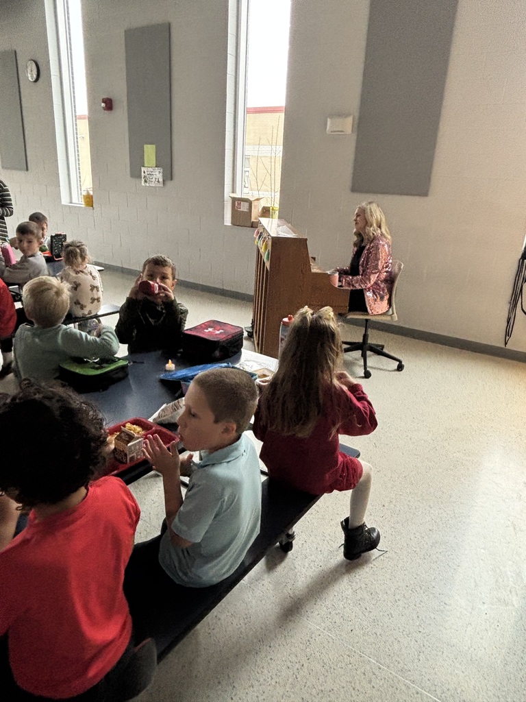A woman wearing a sparkly pink jacket plays the piano in a school cafeteria while young children sit at long lunch tables eating their meals and watching her.