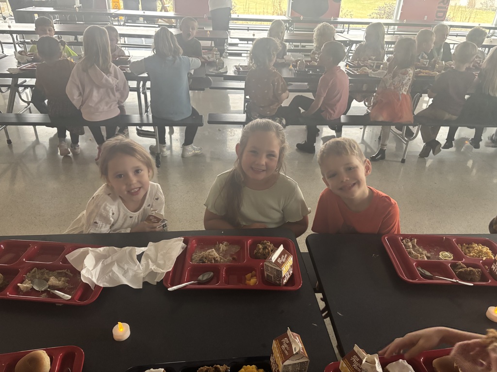 Three young children smile at the camera while eating lunch in a busy school cafeteria. They sit at the edge of a long table with red lunch trays containing a Thanksgiving-style meal, with many other students seated behind them.