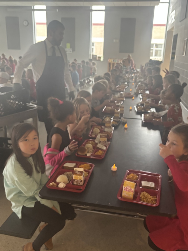 A long cafeteria table is filled with elementary students eating a Thanksgiving-style meal on red trays. A staff member wearing a black apron walks beside the table, and small battery-powered candles line the center of the table.