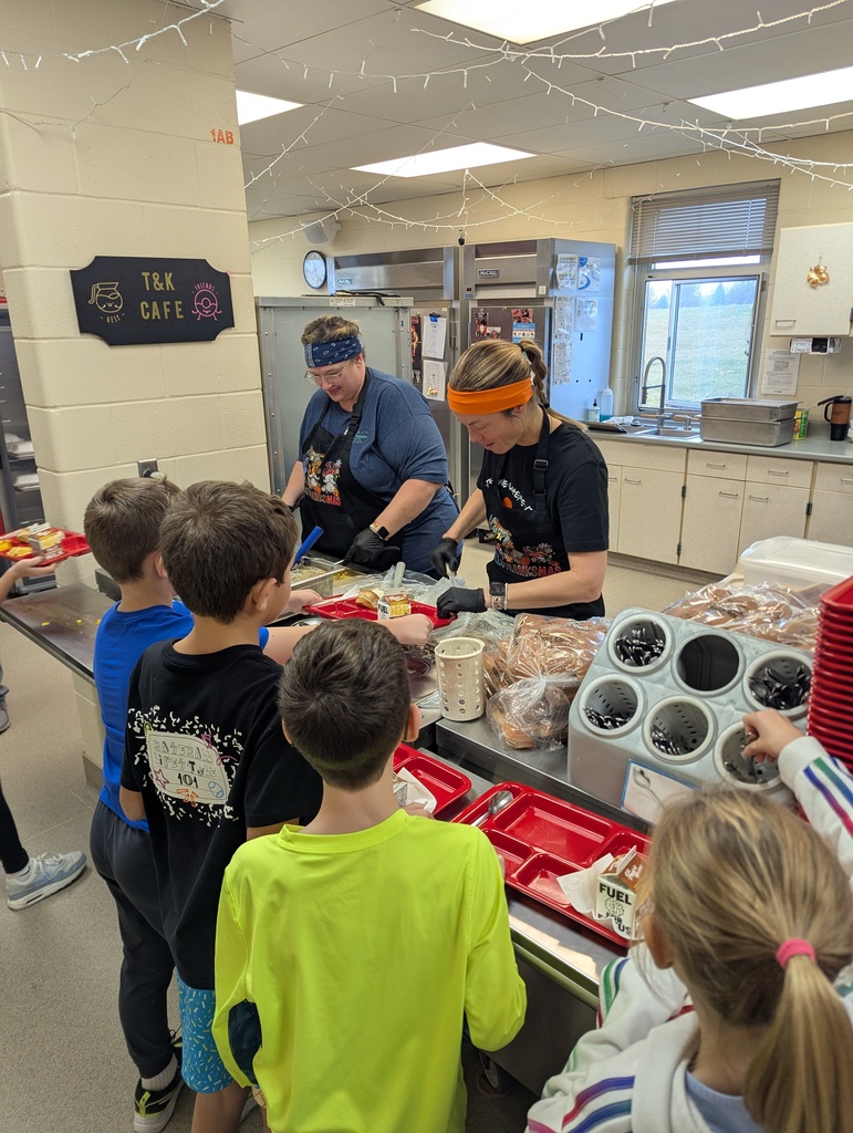 Cafeteria workers serve lunch to a group of elementary students standing in line at the counter. Two staff members wearing aprons, gloves, and headbands are preparing food behind the counter while several children hold red lunch trays.