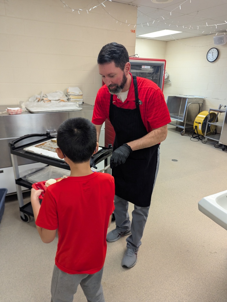 A school staff member wearing a black apron and red shirt serves food to an elementary student holding a red lunch tray.
