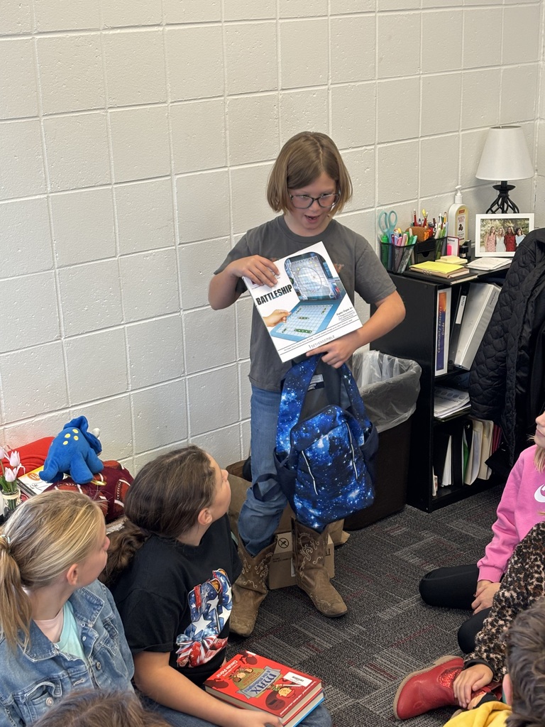 A student stands in front of her classmates holding a Battleship game box in one hand and a blue galaxy-pattern backpack in the other. Several students sit on the floor around her, watching and listening.