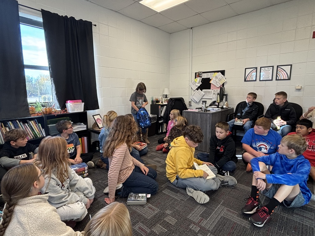 A classroom scene where a group of students sits on the floor in a circle. At the front, the same student stands holding the blue galaxy-pattern backpack while classmates watch.