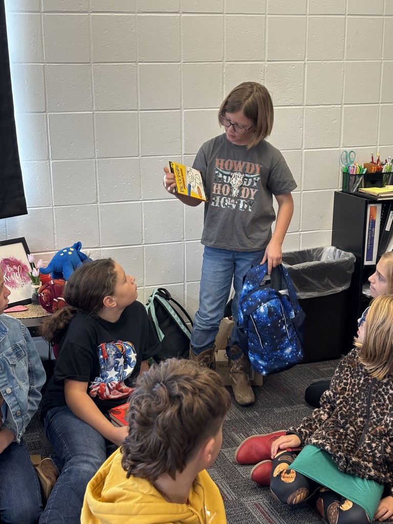 The same student stands in front of her peers, now holding up a book while still holding the blue galaxy-pattern backpack. Students seated on the floor look up at her attentively.