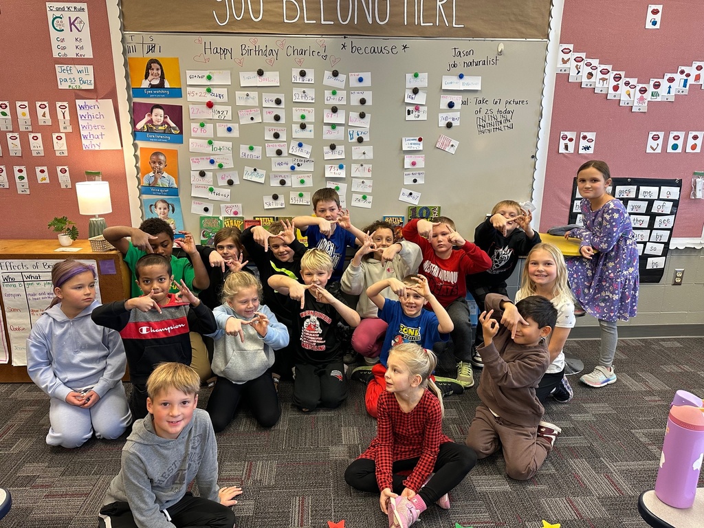 A group of elementary students pose together in a classroom, many of them smiling and forming hand signs with their fingers. They sit and kneel on a carpeted floor in front of a large whiteboard covered with student work, drawings, and math problems.