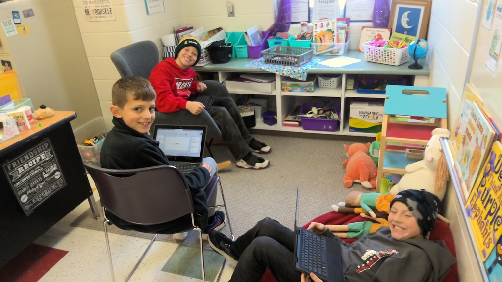 Three elementary students sit together in a cozy classroom corner, each using a laptop. One student sits in a chair smiling at the camera, while the other two sit on cushions and a chair nearby