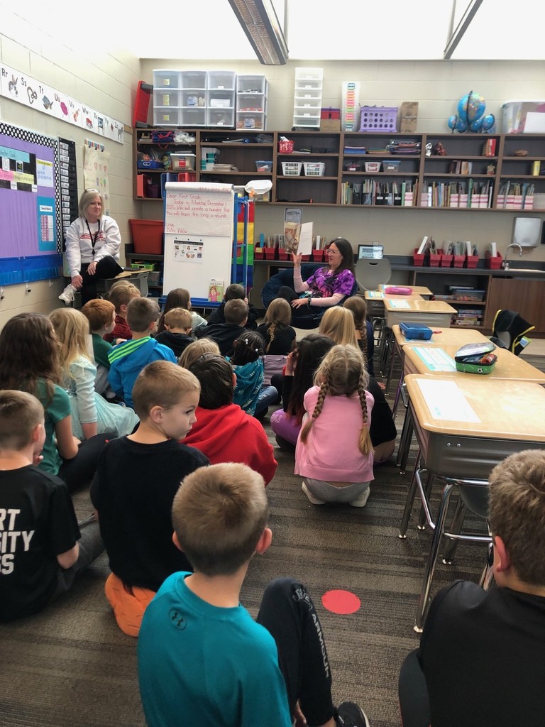 A group of elementary students sit on the floor facing a teacher who is reading a picture book aloud at the front of the classroom. Another adult sits nearby, observing. Behind them are shelves filled with books, bins, and classroom supplies, and a whiteboard with written instructions is positioned beside the teacher.