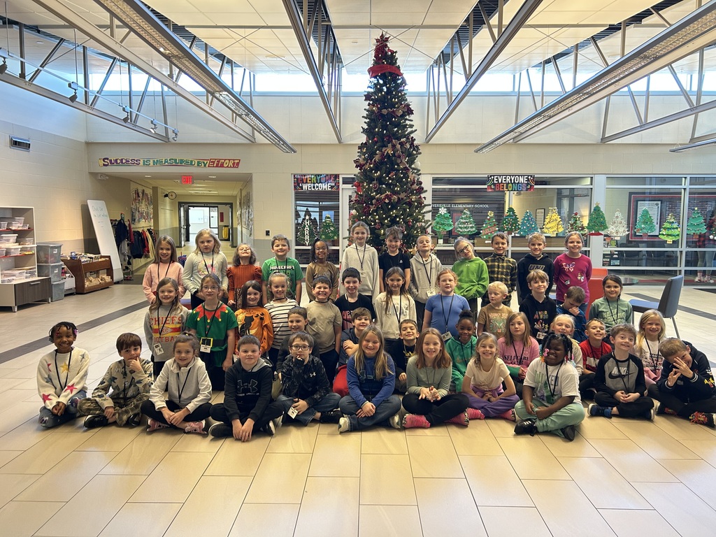 A large group of elementary students pose together in several rows inside a school lobby, smiling at the camera. A tall decorated Christmas tree stands behind them.