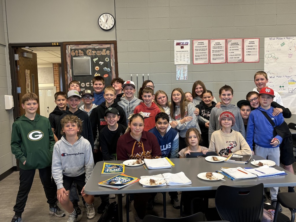 A group of sixth-grade students pose together and smile inside a classroom. Several students sit at a table in front with notebooks and plates of cake, while others stand behind them. Classroom posters and a chalkboard labeled “6th Grade Social Studies” are visible on the wall.