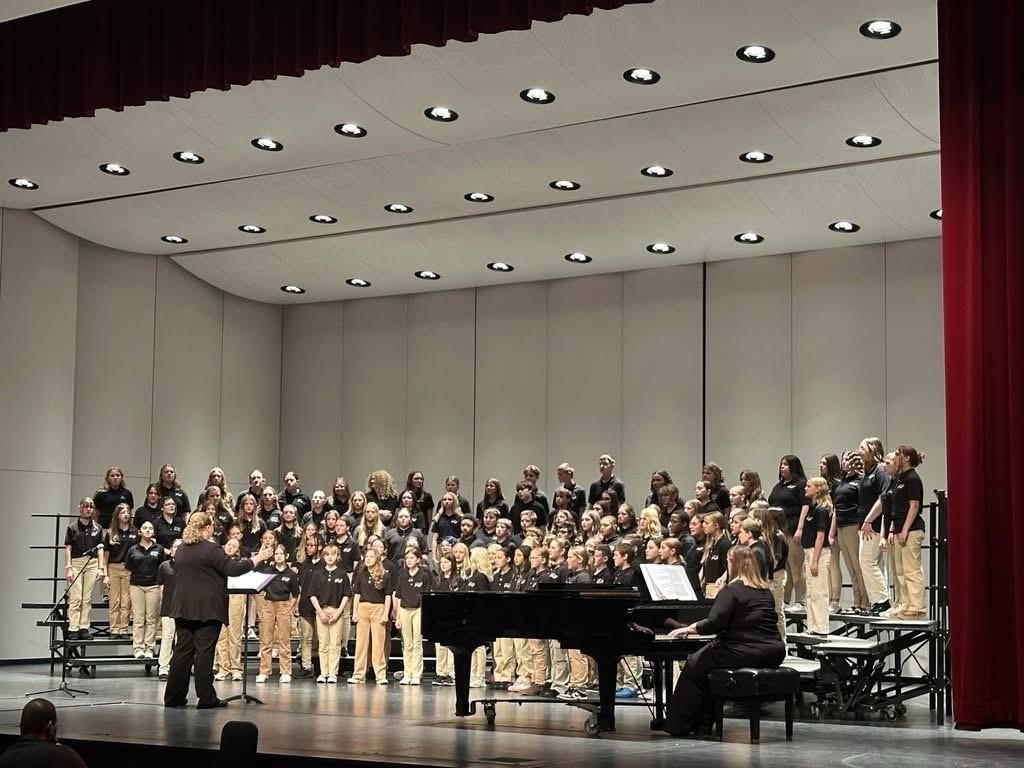 A large middle-school choir stands on tiered risers on a theater stage, wearing matching black shirts and khaki pants. A conductor faces the students at the front left, leading the performance. A pianist sits at a grand piano on the right side of the stage.