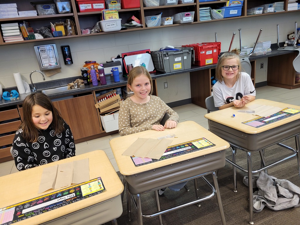 Three girls sit at their desks smiling while working with wheat seeds placed on paper towels.