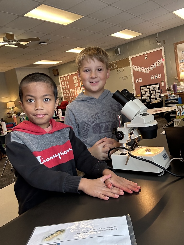 Two young boys smile at the camera while working with a microscope at a classroom table. One boy looks through the eyepiece while the other stands beside him.