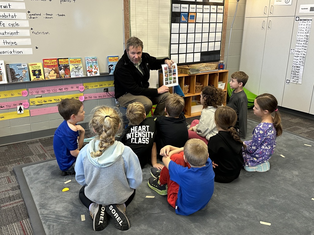 A man sits on the floor at the front of a classroom holding up a laminated sheet with pictures of animals while a small group of elementary students sit in a semicircle on a rug, listening attentively.