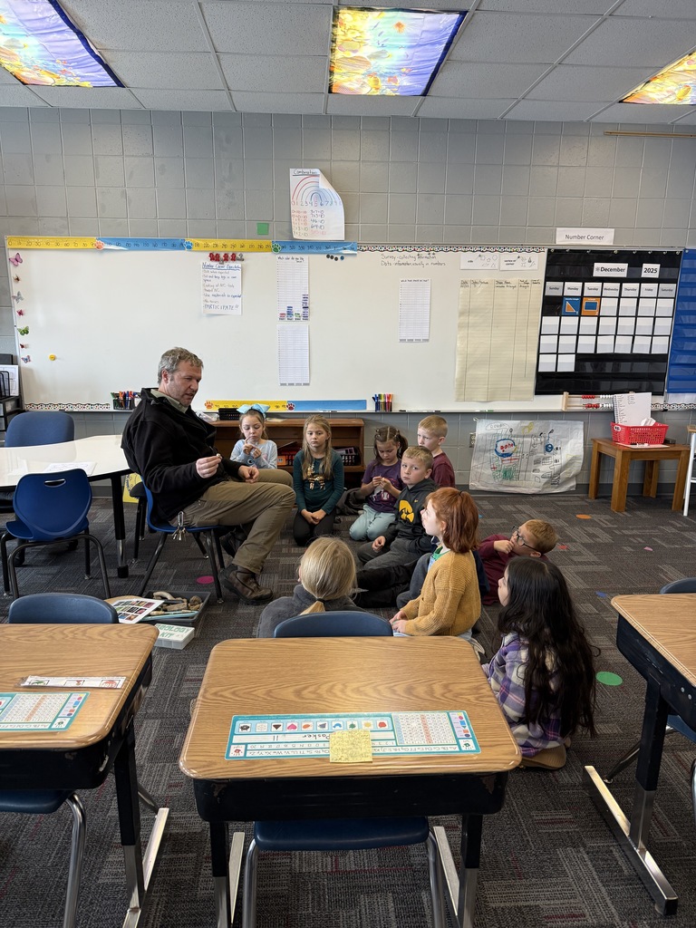 A small group of students sit in a semicircle around a man who is holding a fossil and speaking to them in a classroom.