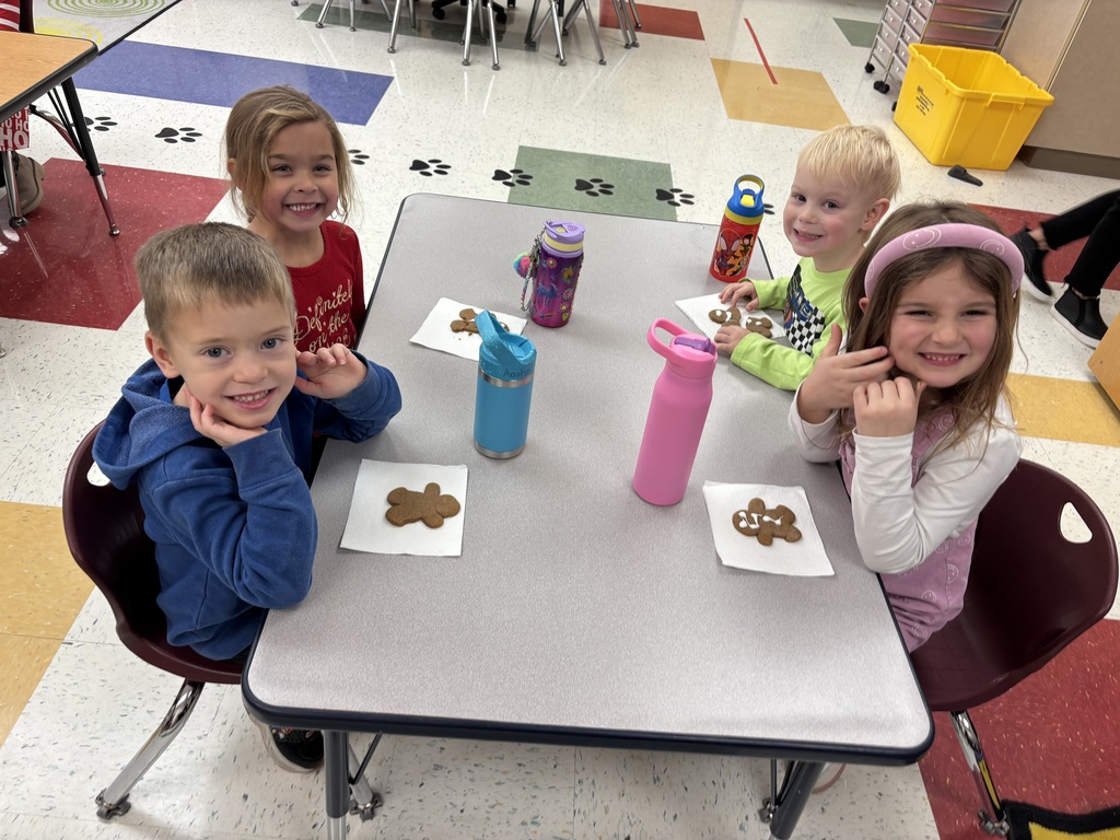 Four young children sit at a table with water bottles and decorated gingerbread cookies on napkins, smiling cheerfully for the camera.