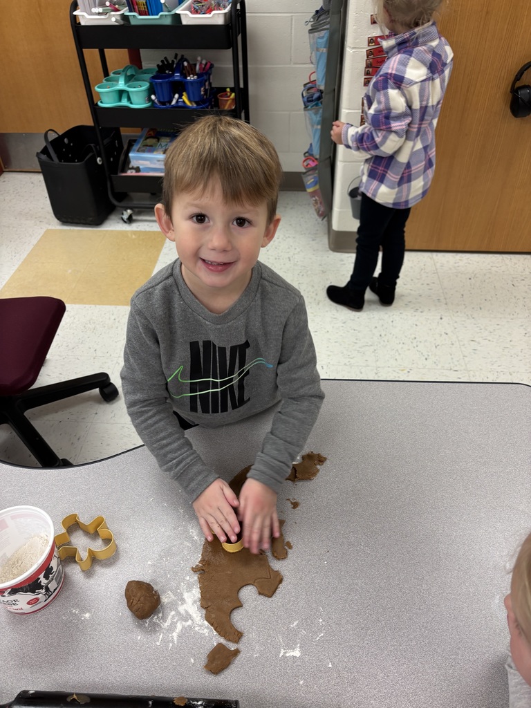 A young boy presses a gingerbread-shaped cookie cutter into dough on a classroom table.
