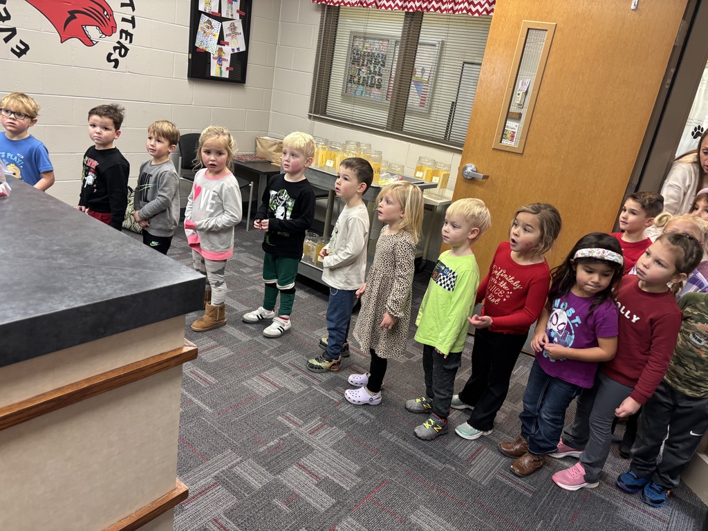 : A group of preschool children stand in a line inside a school hallway, looking toward a counter.