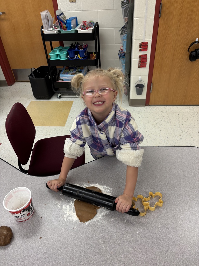 A smiling young girl with glasses and pigtails rolls out gingerbread dough with a rolling pin on a classroom table. Cookie cutters and flour are visible on the table.