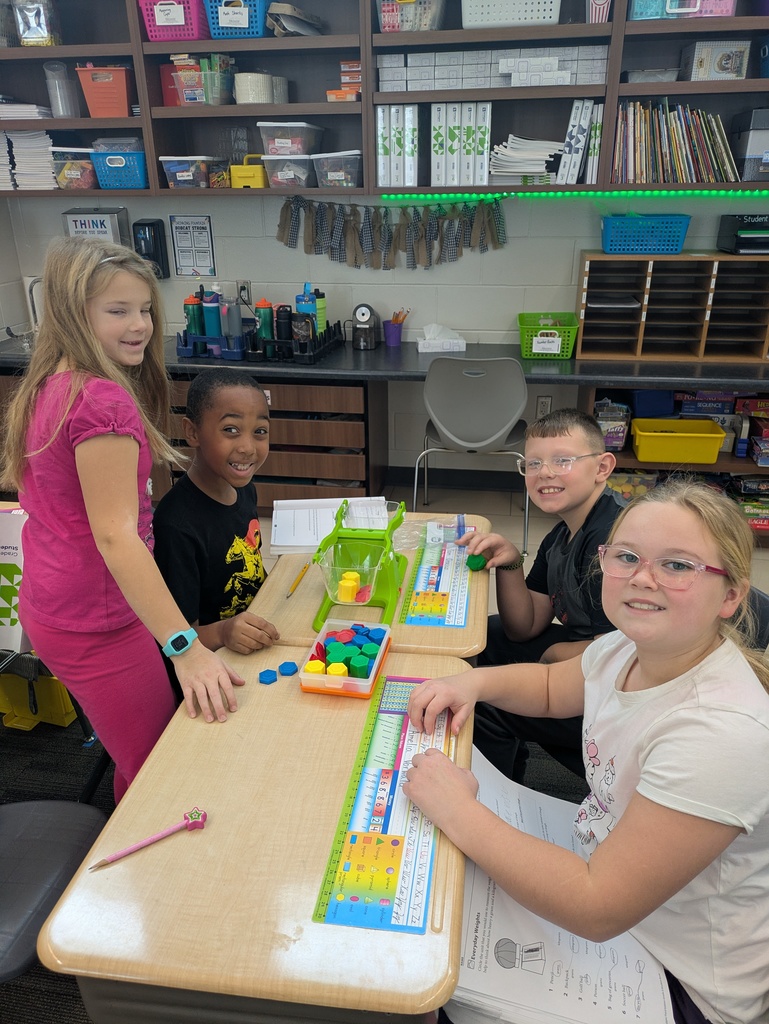 Four students at a desk work with a green balance scale and colorful blocks while smiling at the camera