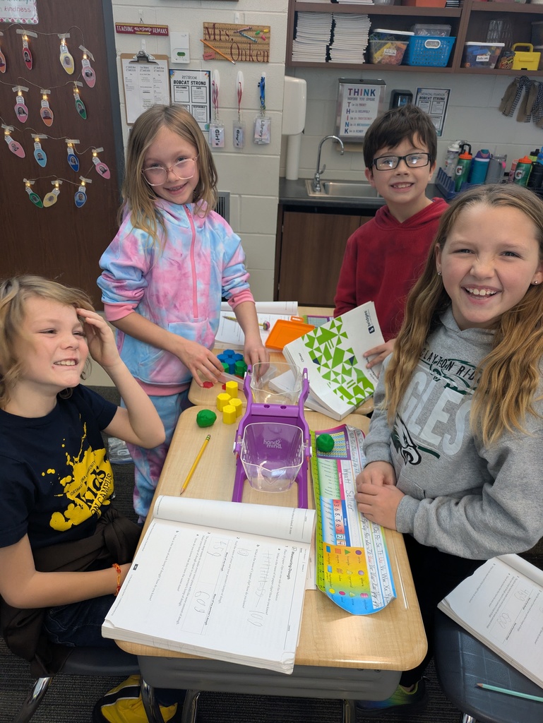 Four students stand and smile while using a purple balance scale and various shapes at a classroom desk with notebooks open.