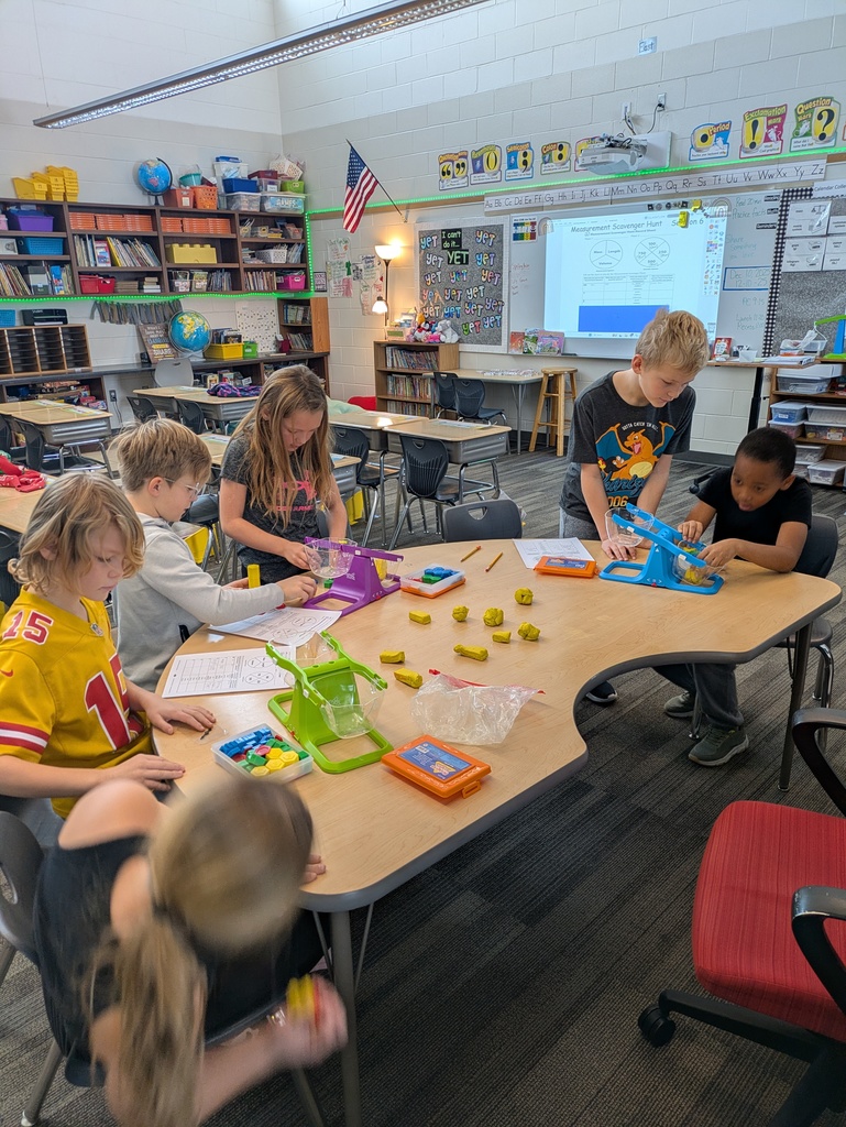 Students gather at a table using balance scales, Play-Doh, and shapes during a math activity, collaborating and writing in their notebooks.