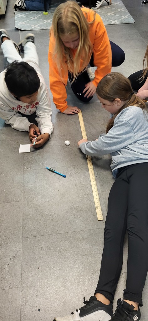Three students work together on a floor activity, using a large wooden ruler to measure the distance a small object has traveled.