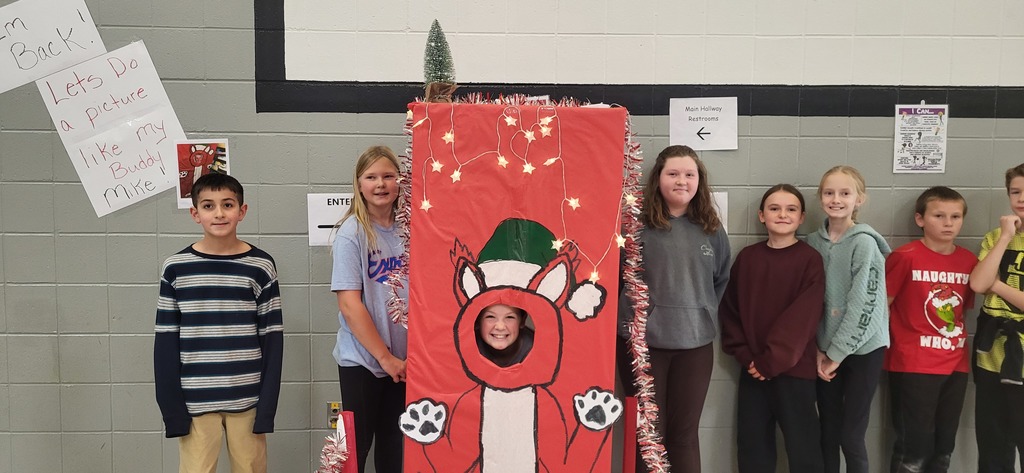 A group of students stand in a hallway next to a red holiday-themed photo booth decorated with lights and a drawing of a reindeer wearing a Santa hat. One student smiles through the face cutout of the booth, while others pose beside it.