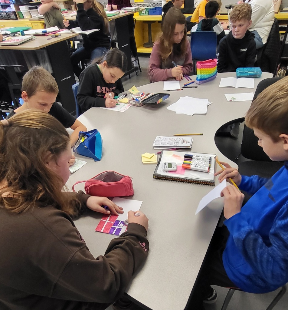 A group of elementary students sit around a classroom table working on drawings or crafts.