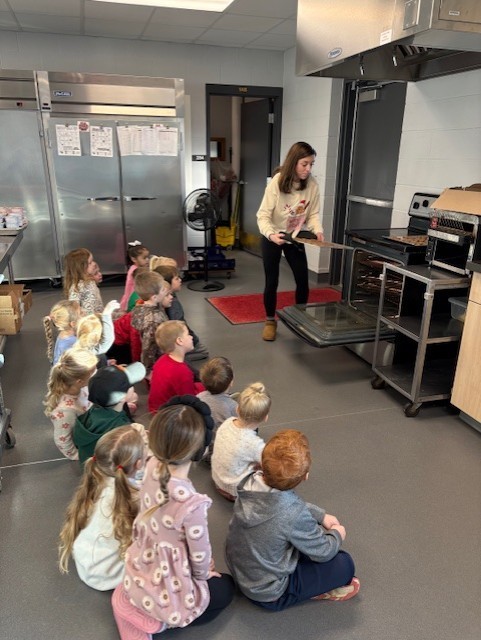 A teacher places a tray into an oven while a group of young children sit on the floor, watching attentively in a school kitchen.