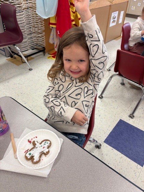 Young students participate in a festive classroom activity, decorating and baking gingerbread cookies using rolling pins, icing, and toppings.
