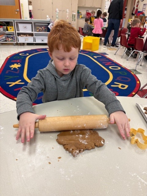 Young students participate in a festive classroom activity, decorating and baking gingerbread cookies using rolling pins, icing, and toppings.