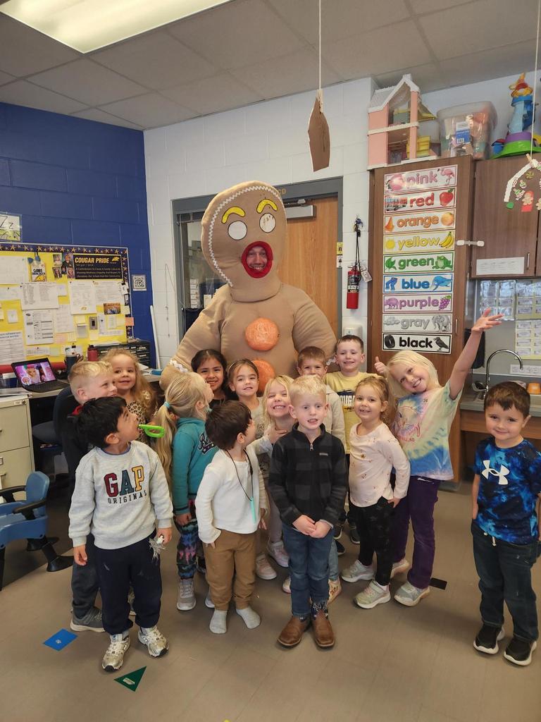 A group of smiling young students poses with an adult dressed in a large gingerbread costume inside a colorful classroom.
