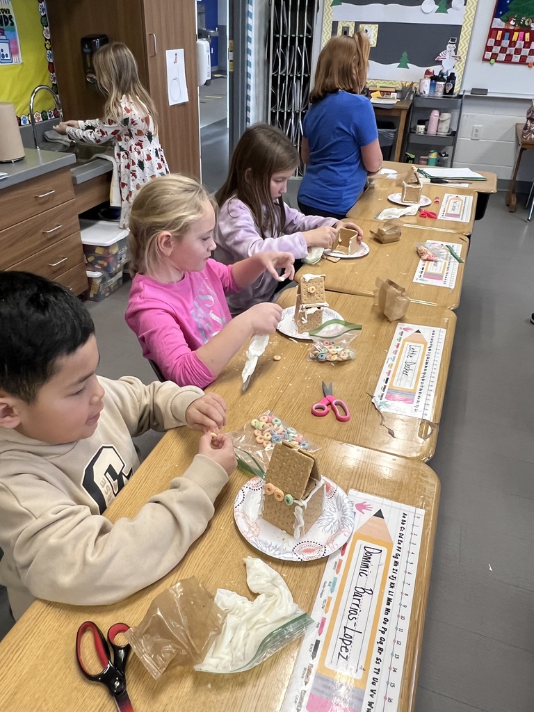Children sit at a classroom table decorating small graham cracker gingerbread houses with frosting and cereal pieces.