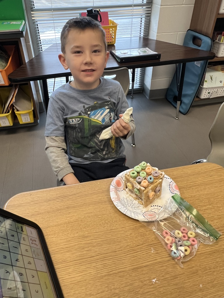 A smiling boy holds a piping bag of frosting next to his finished gingerbread house decorated with colorful cereal.