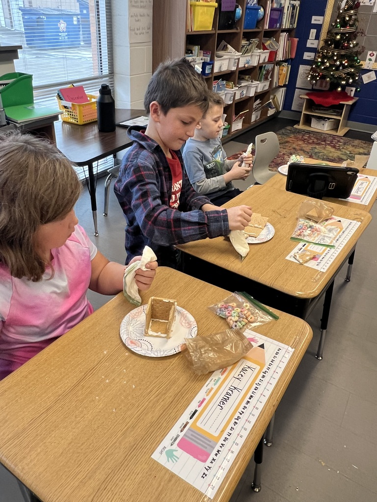 Three children sit at desks decorating graham cracker houses with icing, cereal, and plastic bags filled with frosting.